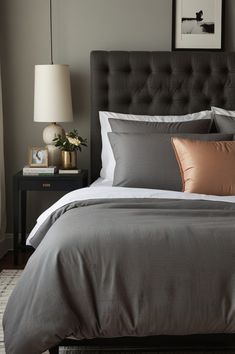 a bed with grey sheets and pillows in a bedroom next to a lamp on a night stand