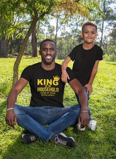 a father and son sitting on the grass in front of a tree wearing matching t - shirts