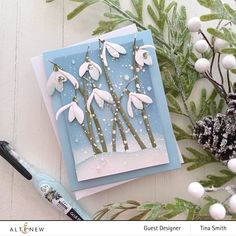 a card with white flowers on it next to some green leaves and snowflakes