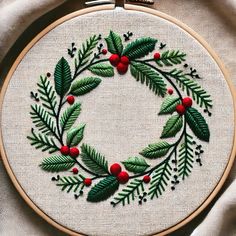 an embroidered christmas wreath with red berries and green leaves is shown on a white cloth