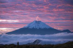 a mountain covered in clouds under a pink and blue sky at sunset with the sun setting behind it