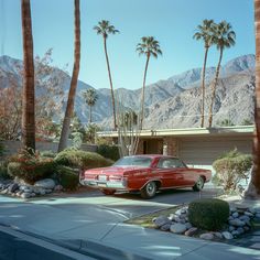 an old red car parked in front of a palm tree lined house with mountains in the background