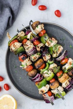 skewered vegetables and meat on a black plate with tomatoes, onions, and lemon wedges