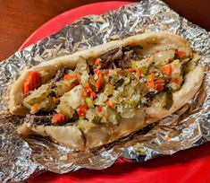 a hot dog covered in lots of toppings on top of aluminum foil with a red plate