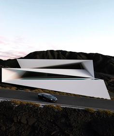 a car driving past a white building on the side of a road with mountains in the background