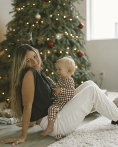 a woman sitting on the floor next to a christmas tree with a baby in her lap