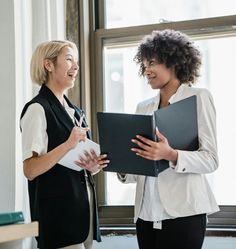 two women standing next to each other in front of a window holding binders and papers