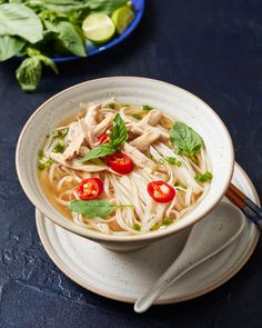 a bowl of soup with noodles, meat and vegetables on a plate next to some basil leaves