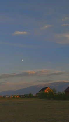 the moon is setting in the sky over a field with houses and mountains behind it