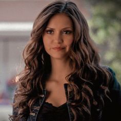 a woman with long brown hair wearing a black shirt and jean jacket looking at the camera