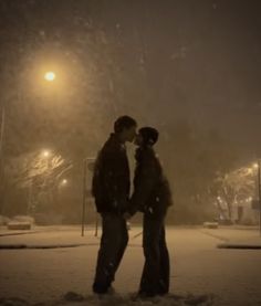 two people standing in the snow at night with street lights behind them and one person kissing the other