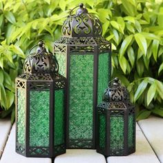 three green lanterns sitting on top of a white tile floor next to bushes and plants