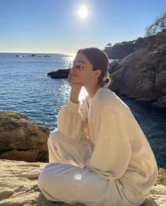 a woman sitting on top of a rock next to the ocean with her hand under her chin