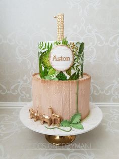 a three tiered cake decorated with greenery and the word ashton on top is sitting on a gold plate