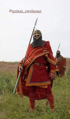 two men dressed in medieval clothing and helmets with swords, standing on the grass field