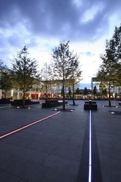 an empty plaza with benches and trees in the background at dusk, lit up by street lights