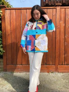 a woman standing in front of a wooden fence wearing a colorful jacket and white pants