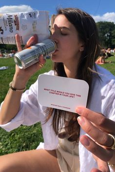 a woman sitting in the grass drinking from a bottle and holding up a card to her mouth