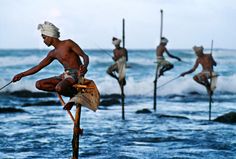 a man standing on stils in the ocean with other men behind him holding poles