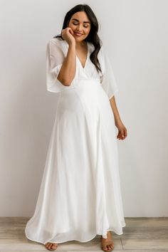 a woman wearing a white dress and holding her hand up to her face while standing in front of a wall