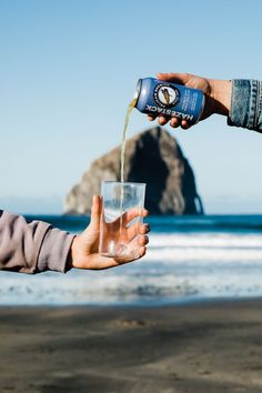 someone is pouring water into a glass on the beach while another person holds out their hand