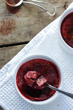 two white bowls filled with red food sitting on top of a wooden table next to spoons