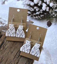 two white bear earrings sitting on top of a piece of wood next to snow covered ground