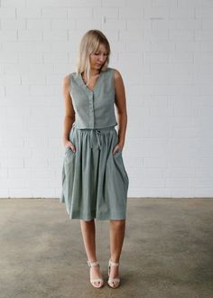 a woman standing in front of a white brick wall wearing a gray dress and sandals