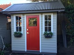 a red door sits in front of a white shed with two windows and potted plants