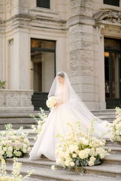 a woman in a wedding dress and veil walking down some steps with flowers on the ground