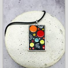 a black and red square pendant with multicolored circles on it sitting on top of a white rock