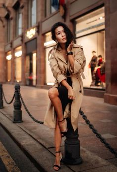 a woman sitting on top of a lamp post in front of a tall building wearing a trench coat