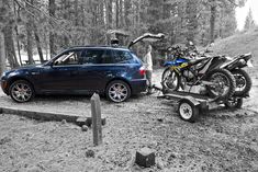 a blue car parked next to a motorcycle on the back of a trailer in front of a forest