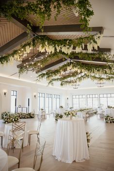 an indoor wedding venue with white tables and greenery hanging from the ceiling, surrounded by tall windows