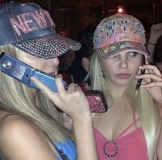 two beautiful young women talking on cell phones in a room with other people and one is holding a cell phone up to her ear