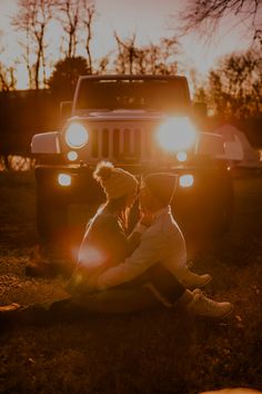 two people sitting on the ground in front of a jeep at sunset with their lights on