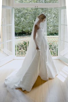 a woman in a white wedding dress is standing by an open window