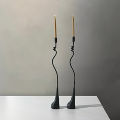 two black candles are standing next to each other on a white table with a gray wall in the background