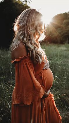 a pregnant woman in an orange dress standing in tall grass with the sun shining behind her