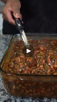 a person using a tong to stir beans in a large glass casserole dish