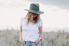 a woman wearing a green hat and white shirt standing in the grass with her hands on her hips