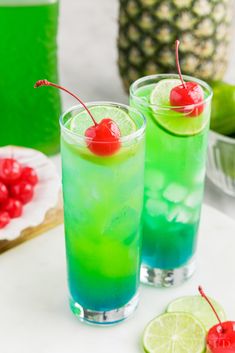 two glasses filled with blue and green liquid next to pineapples, cherries, and lime slices