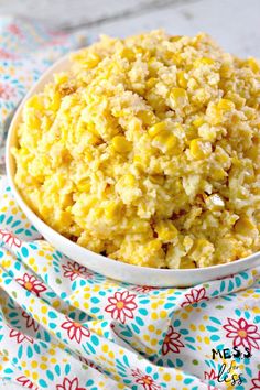 a white bowl filled with corn on top of a colorful table cloth next to a wooden spoon