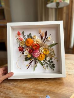 a person holding a white frame with flowers in it on top of a wooden table