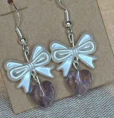 Delicate Bow and Heart Earrings These charming handmade earrings feature a pearlescent white bow charm paired with a translucent purple or pink heart-shaped bead. The bows have a lovely sheen and dimensional appearance, while the faceted heart beads add a touch of sparkle. Details: Sterling silver fish hook earwires for easy wearing Pearlescent white plastic bow charms, approximately 2 cm wide Translucent purple or pink faceted heart beads, approximately 1 cm Total drop length of about 4 cm Perfect for adding a sweet, feminine touch to any outfit. These versatile earrings would make a lovely gift or a special treat for yourself. The soft colors and dainty design make them suitable for both casual and dressier occasions. Each pair is handcrafted with care. Due to the handmade nature, slight Elegant White Jewelry With Pink Bow, Heart-shaped Earrings With Bow For Gift, Cute White Jewelry With Ribbon, Party Jewelry With Pink Bow In White, White Jewelry With Pink Bow For Party, White Party Jewelry With Pink Bow, White Heart Charm Earrings For Party, Heart Shaped Bow Earrings For Party, Party Heart Earrings With Bow