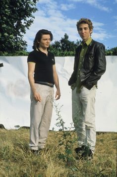 two young men standing next to each other in front of a white wall and trees