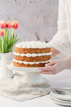 a person holding a cake with frosting and flowers in the background on a table