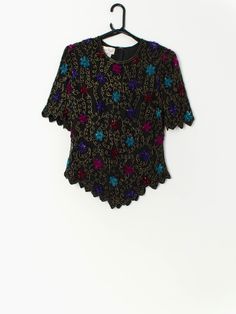 Vintage sequin top in black with floral pattern by Laurence Kazar. This beautiful floral sequin and beaded top features bright blue, pink and purple flowers, short sleeves, shoulder pads, a quarter zip and hook and eye closure on the back. Made from a silk and a polyester lining, this top is the perfect party piece! Our recommended size: Medium Label says: Large Condition: Very good* Material: 100% silk Lining material: 100% polyester  Measurements in inches: Pit to pit: 19 Shoulders: 16 Sleeve: Glamorous Purple Sequin Top, Embellished Short Sleeve T-shirt For Party, Fitted Sequin Party T-shirt, Fitted Sequin T-shirt For Party, Summer Purple Sequined Tops, Fitted Floral Embroidery Blouse For Night Out, Fitted Purple Sequin Top, Embellished Short Sleeve Tops For Party Season, Embellished Short Sleeve Tops For Night Out