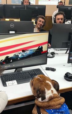 a dog sitting in front of a computer monitor with people looking at the screen behind it