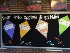 a bulletin board with some kites hanging on it's side and writing that says, soar into spring & sing